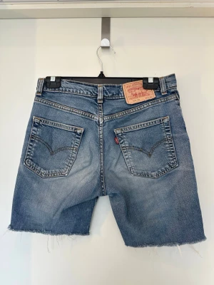 Levi's jeansshorts med rå kant Medium - Klassiska blå jeansshorts från Levi's med rå, fransig kant och femficksmodell. Shortsens denim är i bomull och har en avslappnad passform. Bakfickorna har den ikoniska Levi's-sömmen och läderpatch bak i midjan.
