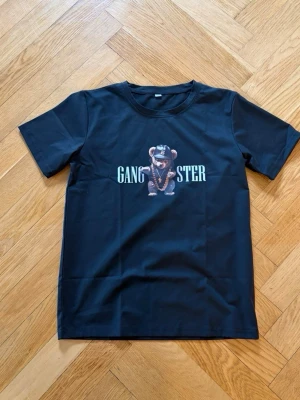 Svart t-shirt med björntryck - Svart t-shirt med coolt tryck av en björn med keps, kedjor och texten 'GANGSTER' på bröstet. Klassisk passform och rund halsringning. Perfekt för dig som vill sticka ut med en streetstyle-look.