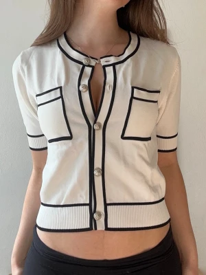 Vit kortärmad kofta med svarta detaljer - Stilren vit kofta med korta ärmar och svarta kontrastkanter. Modellen har rund halsringning, dekorativa fickor framtill och knäppning med silvriga knappar. Ribbstickad nederkant och ärmslut ger en snygg passform. Perfekt för en trendig och chic look.