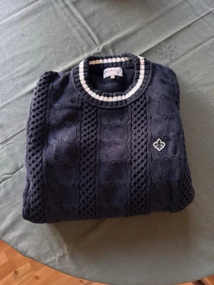 Mörkblå stickad tröja från Morris - Snygg mörkblå stickad tröja från Morris med kabelstickat mönster och vit broderad logga på bröstet. Rund halsringning med vita och grå ränder samt matchande detaljer vid ärmsluten. 