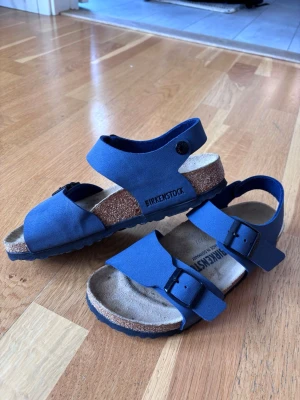 Blå sandaler från Birkenstock - Snygga blå sandaler från Birkenstock med två justerbara remmar och spännen över foten. De har också remmar över hälen för ytterligare komfort. Passar därför utmärkt för barn/ungdom. Sandalerna har en bekväm korksula och ovandel i blått material. Använda sparsamt en sommar. Perfekta för sommaren och ger en cool och avslappnad stil.