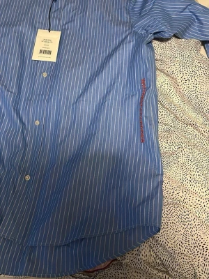 Blå randig skjorta från Whyred - Snygg blå skjorta från Whyred med vita vertikala ränder och klassisk button-down krage. Skjortan har knappar framtill och diskret röd broderad text på sidan. Tillverkad i bomull för en skön känsla och stilren look.
