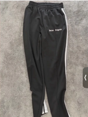 Palm Angels svarta mjukisbyxor - Svarta mjukisbyxor från Palm Angels med vita sidoränder och logga tryckt på vänster lår. Byxorna har resår i midjan och raka ben. Perfekta för en avslappnad streetwear-look.