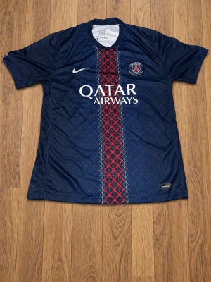 PSG matchtröja Nike blå röd - Snygg blå Paris Saint-Germain matchtröja från Nike med röd och vit rand i mitten, PSG-logga på bröstet och Qatar Airways-tryck. Tillverkad i lätt och ventilerande material, perfekt för fotbollsträning eller supporterliv.