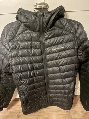 Svart pufferjacka Peak Performance - Snygg svart pufferjacka från Peak Performance med huva och dragkedja framtill. Jackan har quiltad design och diskret logga på bröstet. Perfekt för kalla dagar och riktigt skön att bära. Materialet är lätt och isolerande. Ett hål i vänstra fickan och dragkedjan går inte att dra upp heller på den vänstra fickan. 