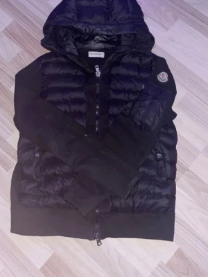 Svart pufferjacka från Moncler - Snygg svart pufferjacka från Moncler med huva och dragkedja framtill. Jackan har quiltade partier på framsidan och ärmar i mjukare material. Klassisk Moncler-logga på ärmen och två fickor med knappstängning. Perfekt för kalla vinterdagar.
