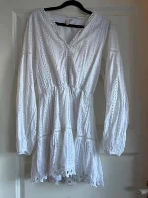 Vit spetsklänning med lång ärm - Söt vit kortklänning med broderad spets och v-ringning. Klänningen har långa ärmar och elastisk midja som ger en fin passform. Detaljerad med volangkant nertill och vackra broderier över hela plagget. Perfekt för sommarens alla tillfällen.