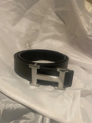 Svart skärp med H-spänne - Stilrent svart skärp med ett ikoniskt H-formad spänne i silver. Skärpet har vita kontrastsömmar längs kanterna och är tillverkat i skinn. Perfekt för att lyfta din outfit med en lyxig touch. 110cm