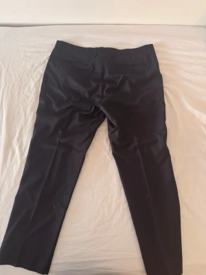 Svarta klassiska kostymbyxor - Snygga svarta kostymbyxor med raka ben och pressveck. Byxorna har bälteshällor, sidofickor och bakfickor med minimalistisk design. Perfekta för en stilren och clean look. Materialet ser ut att vara en slät och mjuk blandning, troligen polyester eller ull.