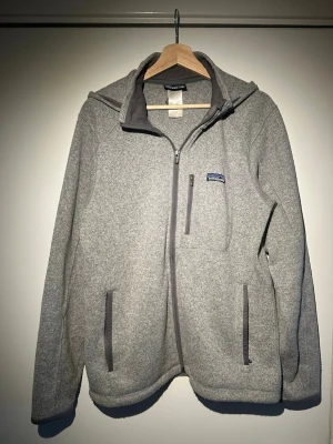 Grå fleece med luva från Patagonia - Mysig grå fleece med luva från Patagonia med dragkedja framtill och två fickor med dragkedja. Tillverkad i mjukt fleecematerial och har en liten logotyp på bröstet. Perfekt för chill dagar eller när du vill ha något bekvämt och stilrent.