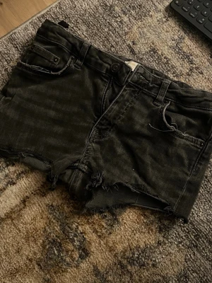 Svarta jeansshorts med fransig kant - Detta är ett par svarta jeansshorts i bomull med råa, fransiga kanter och klassisk femficksdesign. Shortsen har knapp och dragkedja framtill samt bälteshällor. Perfekta för en avslappnad och trendig stil.