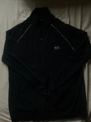 Svart zip-hoodie från BOSS - Snygg svart hoodie från BOSS med dragkedja framtill och diskret logga på bröstet. Har vita detaljer längs axlarna och en klassisk, stilren design. Perfekt för en avslappnad och trendig look.