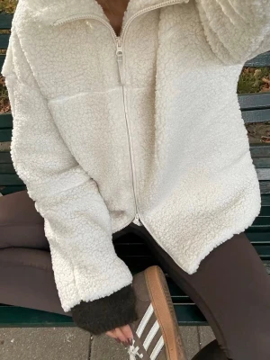 Vit teddyjacka med dragkedja - Mysig vit teddyjacka med hel dragkedja framtill och hög krage. Jackan har en oversized passform och är gjord i fluffigt, mjukt teddy-material som ger en trendig look. Perfekt för våren eller kyligare sommar kvällar.