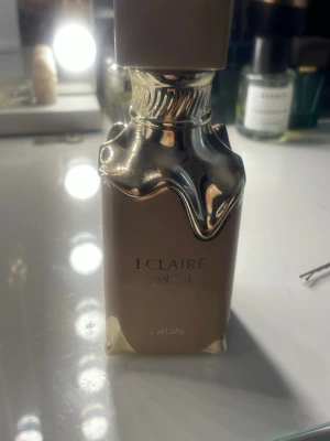 Lattafa Eclaire parfymflaska - Snygg parfymflaska från Lattafa i modellen Eclaire. Flaskan är beige med detaljer i glänsande guld och har en unik design där guldfärgen ser ut att rinna ner över flaskan. Perfekt för dig som gillar lyxiga och trendiga dofter. typ halva kvar