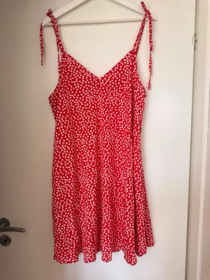 Röd blommig sommarklänning med knytband - Söt röd klänning med vita blommor och smala axelband som knyts upptill. Klänningen har v-ringning och är ärmlös med en lätt utsvängd, kort siluett. Perfekt för varma dagar och har en luftig känsla.