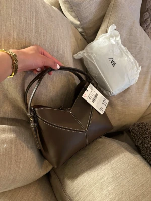 Brun handväska från ZARA - Snygg brun handväska från ZARA i slätt konstläder med vita kontrastsömmar. Väskan har en unik form, guldfärgade metalldetaljer och justerbart axelband. Perfekt för dig som vill ha en trendig accessoar med plats för det viktigaste. Ny oanvänd! Slutsålt på Zaras hemsida💞