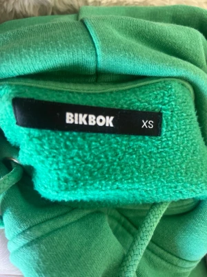 Grön hoodie från BikBok - Snygg grön hoodie från BikBok i storlek XS. Hoodien  är gjord i mjukt, fleecefodrat material som känns riktigt skönt mot huden. Perfekt för dig som gillar en enkel men färgstark stil. Den har ett litet hål i ärmen som ni kan se på bilden🩷