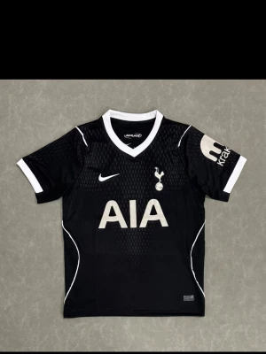 Tottenham Hotspur svart matchtröja Nike - Snygg svart Tottenham Hotspur matchtröja från Nike med vita detaljer och klubbens emblem på bröstet. Tröjan har korta ärmar, V-ringning och stora AIA-loggan framtill. Perfekt för fotbollsträning eller att bära som supporter.