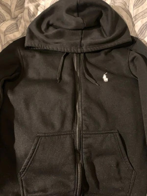Svart hoodie från Polo Ralph Lauren - Svart hoodie med dragkedja från Polo Ralph Lauren. Klassisk design med vit broderad logga på bröstet, känguruficka och dragsko i huvan. Mjuk bomullsblandning som är skön att ha på sig. Perfekt för en avslappnad och stilren look.