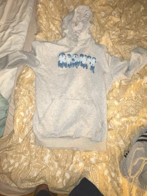 Grå hoodie med blått tryck - Snygg grå hoodie med huva och känguruficka framtill. På bröstet finns ett blått tryck med abstrakt mönster. Hoodien har ribbade muddar vid ärmslut och nederkant. Perfekt för en chill och avslappnad stil.