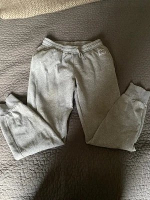 Gråa mjukisbyxor från Andy - Gråa mjukisbyxor från Andy med resår i midjan och dragsko. Byxorna har ribbade muddar vid bensluten och är tillverkade i mjukt bomullsmaterial. Perfekta för en avslappnad och chill stil. Har ett litet hål på höger baksida.