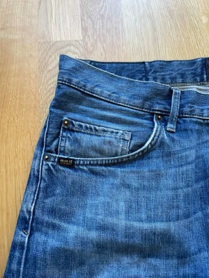 Blå jeans från Tiger of Sweden - Säljer ett par klassiska blå jeans från Tiger of Sweden med raka ben och femficksmodell. Jeansen har snygga slitningar och kontrastsömmar, tillverkade i denim av bomull. Perfekta för en avslappnad och trendig look.