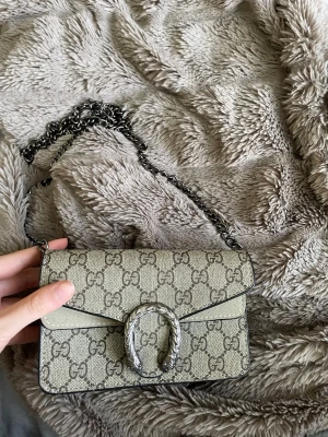 Gucci Dionysus beige handväska - Kan gå ner i pris vid snabb affär. Säljer en beige Gucci Dionysus handväska med klassiskt GG-mönster och silverfärgad kedjerem. Väskan har ett dekorativt spänne framtill och är tillverkad i canvas med detaljer i mocka. Perfekt storlek för det viktigaste och en riktig statement piece.