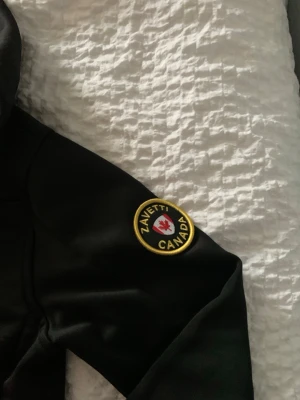 Svart hoodie från Zavetti Canada - Snygg svart hoodie från Zavetti Canada med huva och ett broderat märke på ärmen. Hoodien har en sportig look och detaljer i rött och vitt på insidan av huvan. Perfekt för dig som gillar streetwear och vill ha något stilrent. 