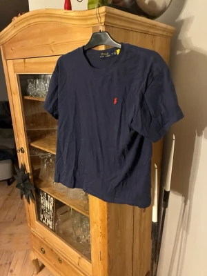 Mörkblå t-shirt från Polo Ralph Lauren - Klassisk mörkblå t-shirt från Polo Ralph Lauren med den ikoniska röda logon broderad på bröstet. T-shirten har rund halsringning och korta ärmar. Tillverkad i mjuk bomull för en skön och avslappnad stil. Använd ett fåtal gånger och har storleken M