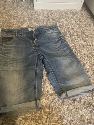 Blå jeansshorts med upprullade ben - Säljer ett par klassiska blå jeansshorts från Levi's med upprullade benslut. Shortsen har fem fickor, knappgylf och en avslappnad passform. Perfekta för varma dagar och enkel att matcha med t-shirt eller hoodie.