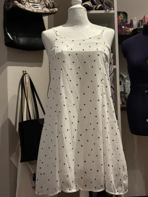 Prickig Sommarklänning - Söt och stilren vit klänning med svarta prickar i klassisk polka dot-stil. Klänningen har tunna axelband och en luftig passform som gör den perfekt för varma sommardagar. Den enkla men tidlösa designen gör den lätt att styla både till vardags med sneakers eller mer uppklätt med sandaler/klackar. Funkar även för mindre storlekar.