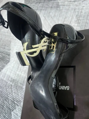 Svarta YSL Opyum pumps i lackläder ej äkta  - Säljer ett par svarta Opyum pumps från Saint Laurent i glansigt lackläder. Skorna har en ikonisk YSL-logga i guld som klack, öppen tå och smal ankelrem. Perfekta för dig som vill sticka ut med en lyxig och trendig look. Ej äkta 