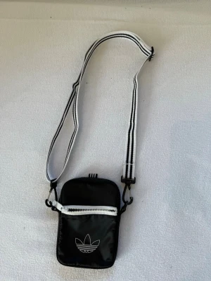 Svart axelväska från Adidas - Snygg svart axelväska från Adidas med vit dragkedja och klassiska trefoil-loggan framtill. Väskan har justerbart axelband i svartvitt och är tillverkad i slitstarkt syntetmaterial. Perfekt för att bära det viktigaste med stil.