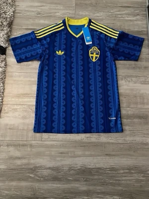 SVERIGE OFFICIELL BORTA TRÖJA VM 2026 - Stilren och unik design i blått med gula detaljer – framtagen i klassisk Adidas-stil med ikoniska tre stripes och exklusiva detaljer. Inspirerad av svensk tradition och modern fotbollskultur. Perfekt för dig som vill sticka ut och stötta Sverige under VM 2026.  ✔️ Bekväm passform ✔️ Broderade märken ✔️ Snygg design ✔️ Perfekt för match, samling eller vardag  Visa ditt stöd för Sverige med den officiella VM-tröjan 2026