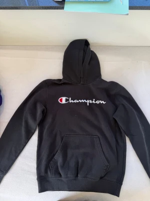 Svart Champion hoodie med logga - Snygg svart hoodie från Champion med stor vit och röd logga broderad på bröstet. Klassisk känguruficka framtill och mjuk insida. Perfekt för en avslappnad och sportig stil. Tillverkad i bomullsblandning och har en rejäl huva.