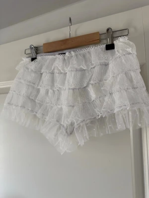 Vita spetsiga pyjamasshorts från S - Supersöta vita pyjamasshorts från Emma och Malena med flera lager volangig spets och liten rosett framtill. Shortsen har elastisk midja och är gjorda i ett tunt, luftigt material som känns lyxigt och romantiskt. Perfekta för mysiga hemmakvällar.