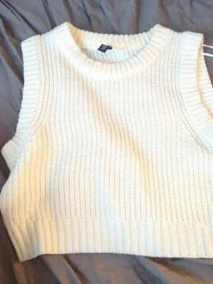 Vit stickad väst från H&M - Säljer en vit stickad väst från H&M med bred ribbad kant vid hals och ärmhål. Västen har en klassisk, enkel design och är gjord i mjukt stickat material. Perfekt att styla över en skjorta eller t-shirt för en trendig lager-på-lager-look.