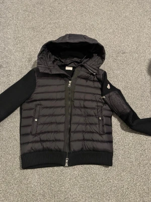 Svart cardigan från Moncler - Säljer en svart cardigan från Moncler med stickade ärmar och huva. Jackan är i storlek L men passar perfekt i storlek M. Jackan har två fickor med knappar, ribbade muddar och ett Moncler-märke på ena ärmen. Materialet är en kombination av quiltad nylon och stickad textil. Perfekt för kalla vinterdagar. 