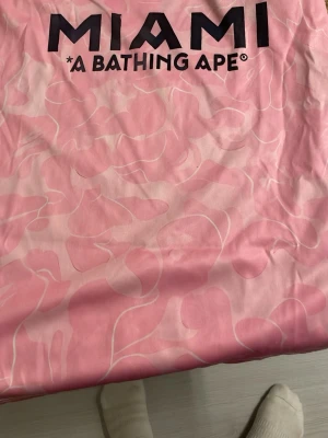 Rosa Miami t-shirt från A Bathing Ape - Säljer en rosa t-shirt från A Bathing Ape med MIAMI-tryck och ikonisk ape-logo på bröstet. T-shirten har ett unikt rosa camouflage-mönster och är tillverkad i mjuk bomull. Perfekt för dig som vill sticka ut med streetwear-vibes.