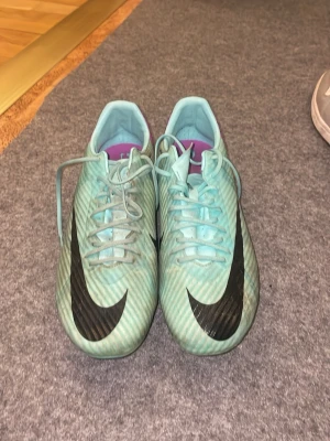 Nike Air Zoom turkosa fotbollsskor - Säljer ett par Nike Air Zoom fotbollsskor i turkos med svarta detaljer och lila häl. Skorna har snörning, stora svarta Nike-loggor och dobbar för spel på gräs. Snygg design och lätta material som ger bra grepp på planen.