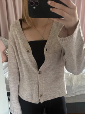 Beige kofta med knappar - Mysig beige kofta med v-ringning och tre stora knappar framtill. Koftan har långa ärmar och är stickad i ett mjukt material, perfekt för lager på lager. Enkel och stilren design som passar till det mesta.