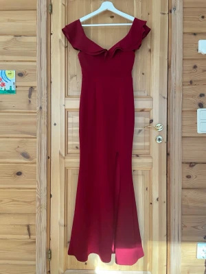 Vinröd långklänning med volang - Elegant vinröd långklänning med offshoulder och volangdetalj vid axlarna. Klänningen har en slits framtill och en figurnära passform som framhäver siluetten. Perfekt för speciella tillfällen där du vill sticka ut. Använd en gång som balklänning!❤️