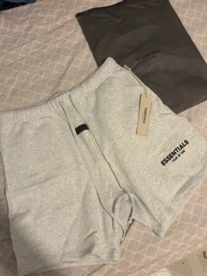 Essentials Fear of God shorts grå - Snygga ljusgrå shorts från Essentials Fear of God med svart logga på benet. Modellen har elastisk midja med snörning och är tillverkad i mjuk bomull, perfekt för en avslappnad stil. Kommer med originalpåse och tag kvar.