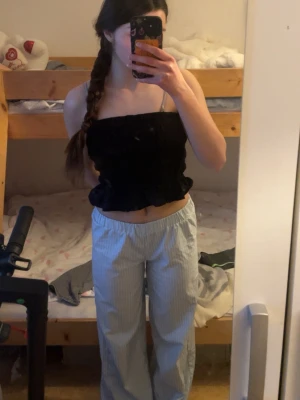 Svart bandeau tubtopp med volang - Svart bandeau tubtopp med smockad struktur och volangkant upptill och nedtill. Toppen är ärmlös och har en croppad passform som sitter tight mot kroppen. Perfekt för en trendig och avslappnad stil.