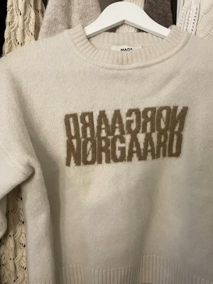 Vit stickad tröja Mads Nørgaard - Snygg vit stickad tröja från Mads Nørgaard med beige logotyp framtill. Tröjan har rund halsringning och ribbade muddar vid ärmslut och nederkant.
