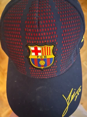 FC Barcelona keps med Messi-signatur - Snygg marinblå keps med röda mesh-detaljer och FC Barcelona-logga framtill. Kepsen har gul Messi-signatur och nummer 10 broderat på sidan. Perfekt för dig som älskar fotboll och vill visa ditt stöd för Barca. Justerbar passform och skön att bära.