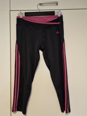 Svarta Adidas träningstights med rosa detaljer - Säljer ett par svarta träningstights från Adidas med rosa ränder längs sidorna och rosa linning. Tightsen är i stretchigt funktionsmaterial och har en kroppsnära passform, perfekta för gym eller löpning.