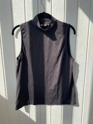 Mörkgrå polotopp från YAYA Women - Snygg mörkgrå ärmlös polotopp från YAYA Women. Toppen har en hög krage och är tillverkad i mjuk bomull, perfekt för en clean och stilren look. Passar till många olika outfits och är enkel att matcha.