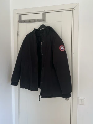 Svart dunjacka från Canada Goose - Svart dunjacka från Canada Goose med huva och klassisk logotyp på ärmen finns ett litet hål inne jackan inget som påverkar utsidan 