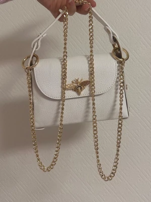 Vit handväska med guldkedja och bi-detalj - Snygg vit handväska i läderimitation med guldfärgad kedja och dekorativt bi i metall på locket. Väskan har både axelrem i kedja och ett kortare handtag. Perfekt accessoar för att lyfta din outfit.